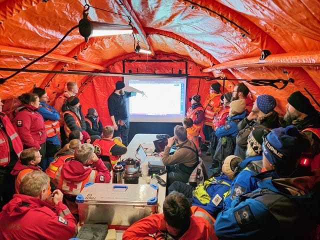 En gruppe med frivillige redningsmannskaper og politi sitter rundt et bord i et telt og ser på en stor skjerm.