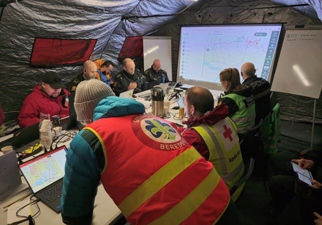 En gruppe med frivillige redningsmannskaper og politi sitter rundt et bord i et telt og ser på en stor skjerm.