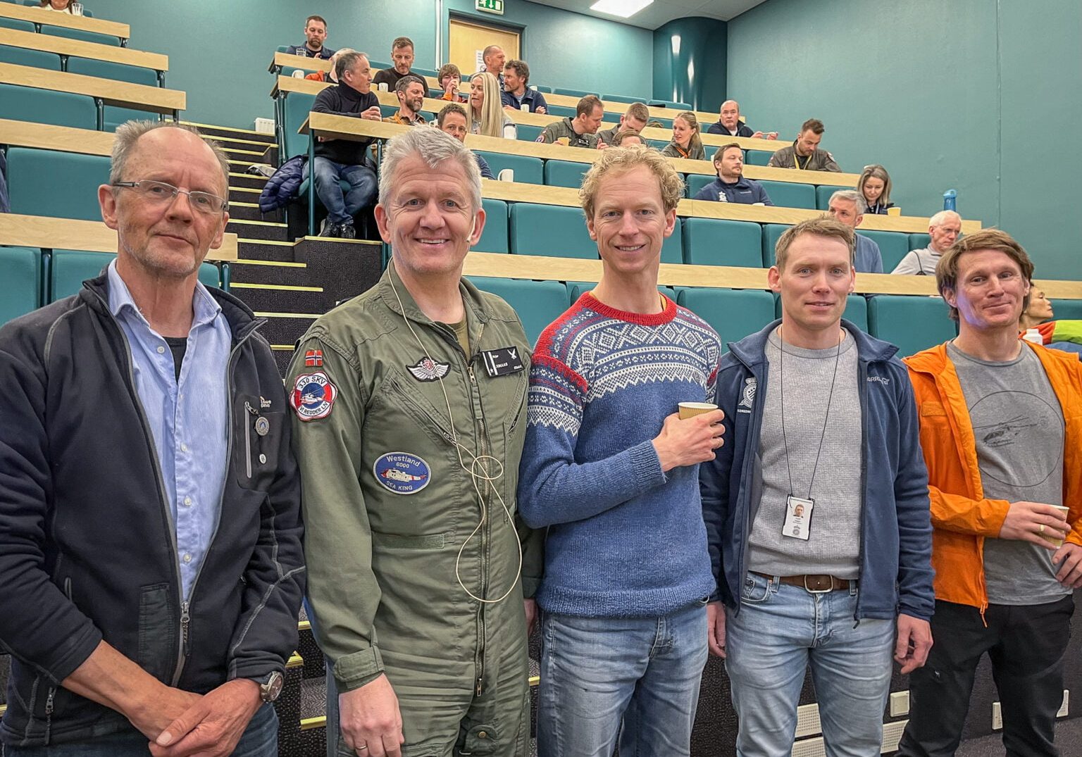 Fem menn står på rekke foran et auditorium,