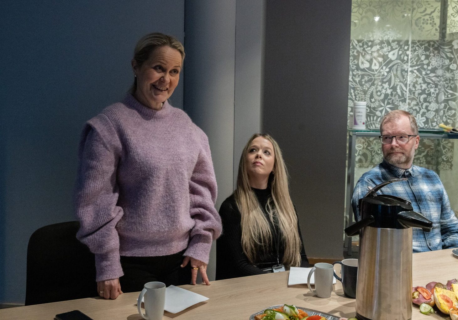 En kvinne står ved et bord der det også sitter to andre mennesker.