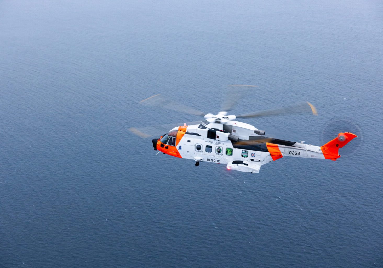 Bilde av et redningshelikopter over sjø, tatt fra et annet helikopter som flyr litt skrått over det første helikopteret.