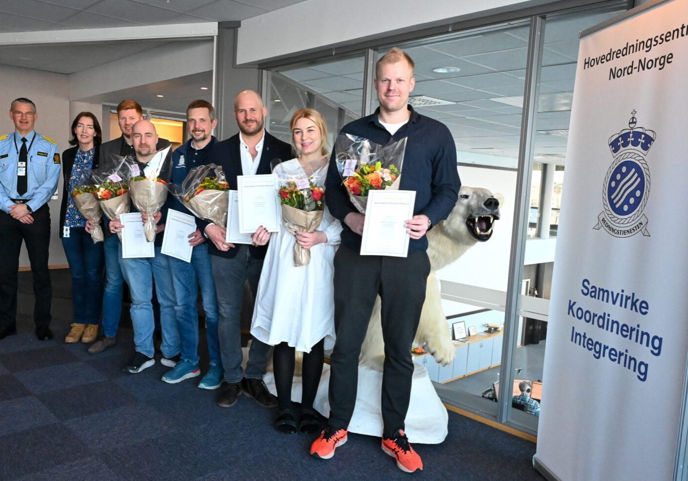 En gruppe mennesker står på rekke og rad og holder diplomer og blomsterbuketter