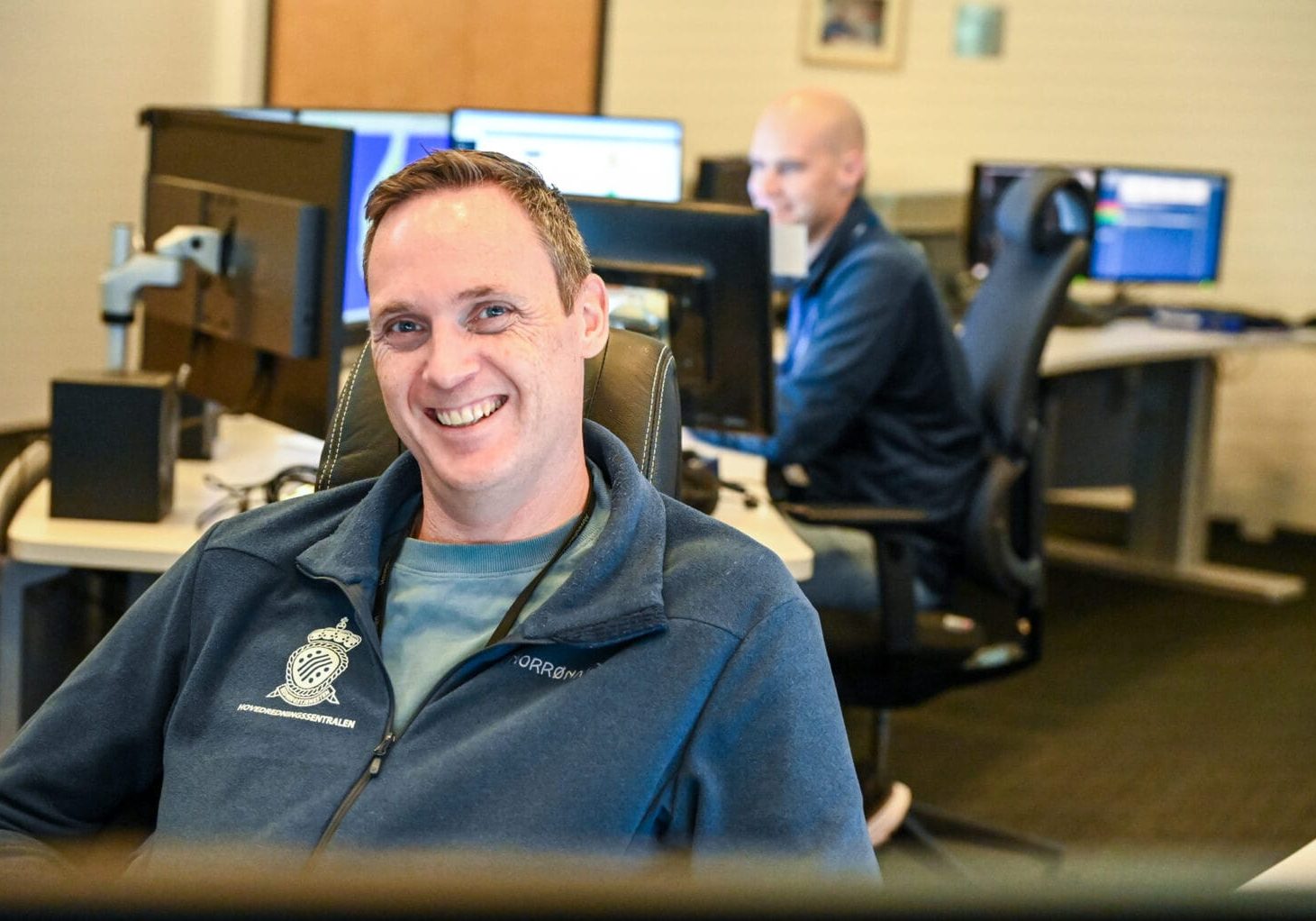 Mann smiler, sitter innendørs, annen mann er uskarp i bakgrunnen