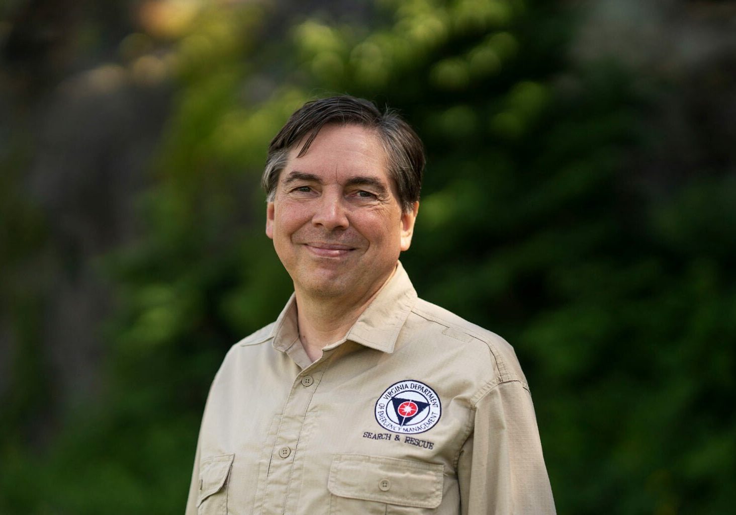 Mann med lys skjorte smiler til fotografen. Skog i bakgrunnen.